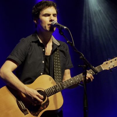 Robert standing on stage with his guitar in a dark grey short sleeve jacket and a dark blue stage background under stage lights.