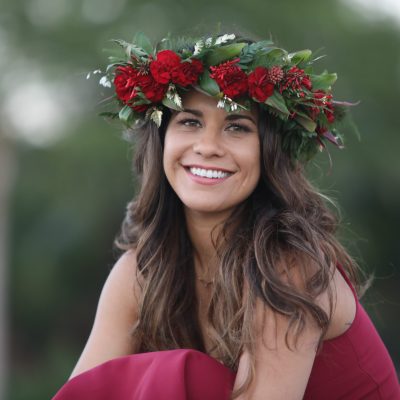 Picture of Kimie Miner in a beautiful maroon dress wearing a dark pink rose headdress with long brown hair, smiling, against a green tree background that is out of focus.